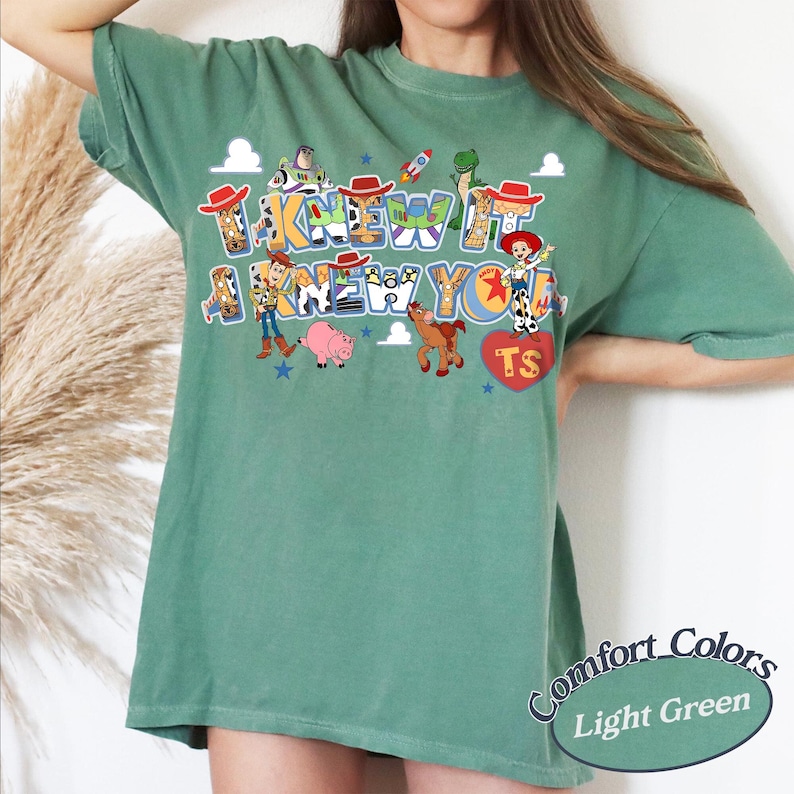May include: A light green short-sleeve t-shirt featuring a graphic print of Toy Story characters including Woody, Buzz Lightyear, Jessie, Rex, Hamm, and Bullseye. The text reads I KNEW IT I KNEW YOU with a red heart containing the letters TS.