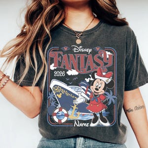 May include: Dark gray t-shirt featuring a Disney Fantasy cruise ship design. The graphic includes Minnie Mouse in a sailor outfit, the text "Disney Fantasy 2026", and a space for a name. The design is framed with red and blue borders.