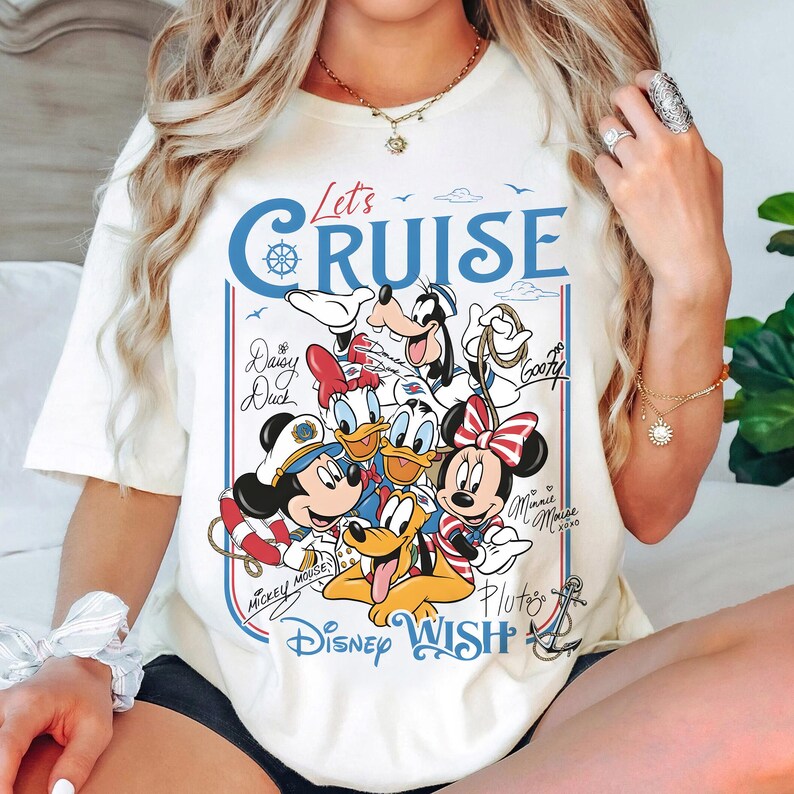 May include: Cream-colored t-shirt featuring a graphic of Disney characters, including Mickey Mouse, Minnie Mouse, Donald Duck, and Goofy. The design includes the text "Let's Cruise" and "Disney Wish" in blue and red.