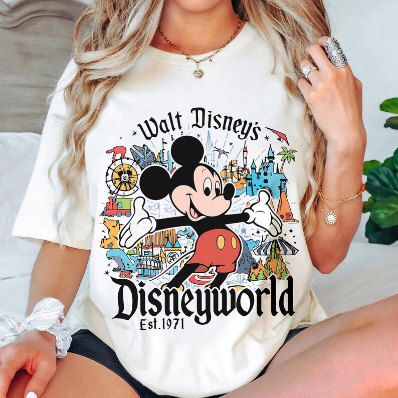 May include: Cream-colored t-shirt featuring Mickey Mouse with the text "Walt Disney's Disneyworld Est. 1971." The graphic includes colorful illustrations of Disney park attractions. The shirt has short sleeves.