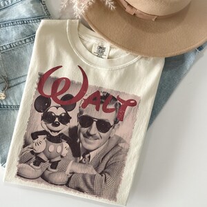 May include: Cream-colored t-shirt featuring a vintage-style graphic of Mickey Mouse and Walt Disney wearing sunglasses, with the word "Walt" in burgundy script. The shirt is folded on a pair of blue jeans, with a beige hat and necklace in the background.