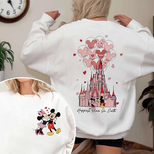 May include: White sweatshirt featuring a graphic of Mickey and Minnie Mouse kissing. The back of the sweatshirt has a castle design with Mickey Mouse balloons and the words "Happiest Place On Earth."