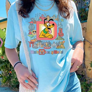 May include: Light blue t-shirt featuring a colorful graphic of Pluto from Disney World. The graphic includes the text "Walt Disney World" and various park landmarks. The shirt is a relaxed fit.