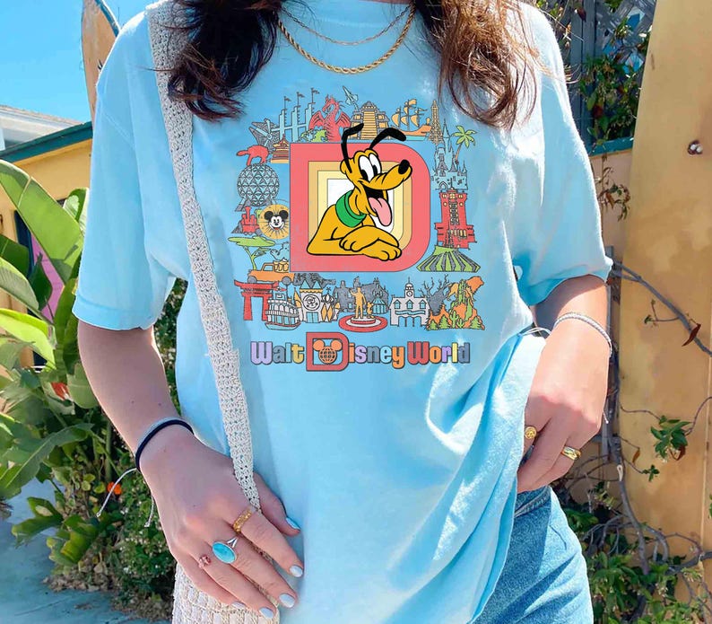 May include: Light blue t-shirt featuring a colorful graphic of Pluto from Disney World. The graphic includes the text "Walt Disney World" and various park landmarks. The shirt is a relaxed fit.