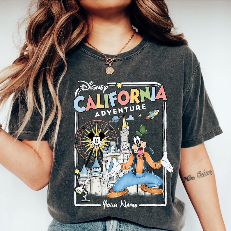 Puede incluir: Camiseta gris oscuro con un gr&aacute;fico colorido de Disney's California Adventure. El dise&ntilde;o incluye a Mickey Mouse en una noria, Goofy, un castillo y el texto "California Adventure". La camiseta tiene un aspecto vintage y desgastado.