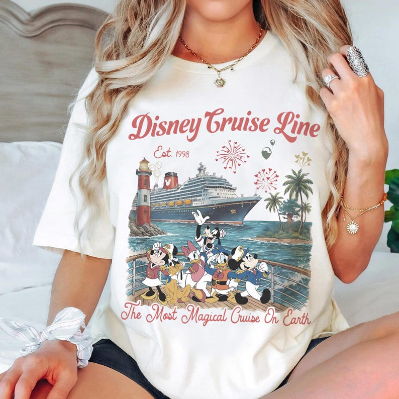 May include: Cream-coloured t-shirt with a Disney Cruise Line graphic. The design shows a cruise ship, lighthouse, palm trees, fireworks, and Disney characters. Text reads "Disney Cruise Line Est. 1998" and "The Most Magical Cruise On Earth."