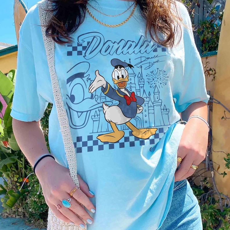 May include: Light blue t-shirt featuring a graphic of Donald Duck in a navy blue jacket and red bow tie, with the text "Donald" above the character. The design includes a castle and fireworks. The shirt has short sleeves.