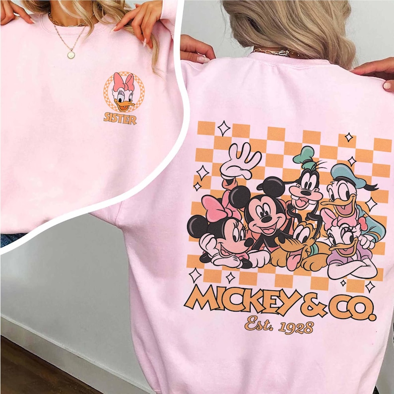 May include: Pink sweatshirt with a checkered pattern and a graphic of Mickey Mouse and friends. The text "Mickey & Co. Est. 1928" is printed below the graphic. The front of the sweatshirt has a smaller graphic of Daisy Duck with the word "Sister" printed below it.