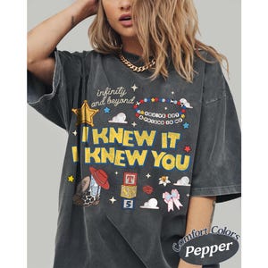May include: A charcoal grey short-sleeve t-shirt featuring a graphic print with the text "infinity and beyond," "YOU'VE GOT A FRIEND IN ME," and "I KNEW IT I KNEW YOU." The design includes a cowboy boot, alphabet blocks, a star, and a bow.