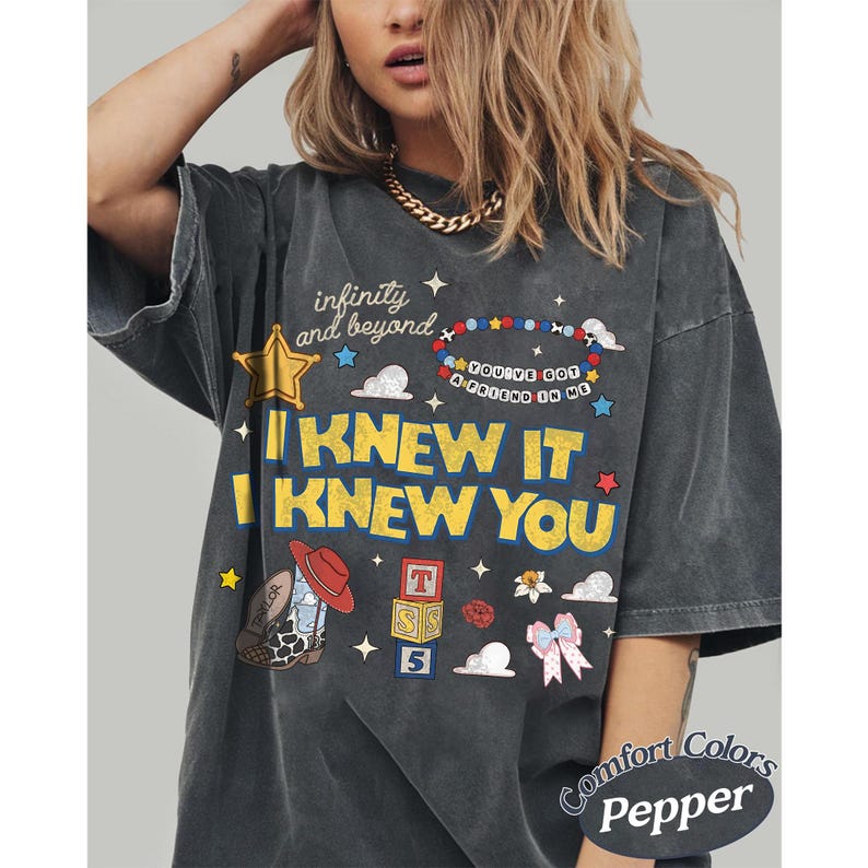May include: A charcoal grey short-sleeve t-shirt featuring a graphic print with the text "infinity and beyond," "YOU'VE GOT A FRIEND IN ME," and "I KNEW IT I KNEW YOU." The design includes a cowboy boot, alphabet blocks, a star, and a bow.