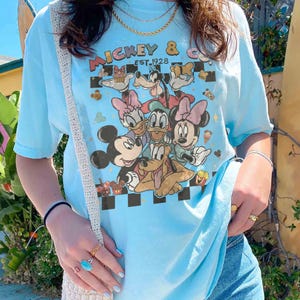 May include: Light blue t-shirt featuring a graphic of Mickey Mouse and friends. The design includes Mickey, Minnie, Donald, Daisy, and Goofy, with the text "Mickey & Co. EST. 1928". The shirt has short sleeves and a crew neck.