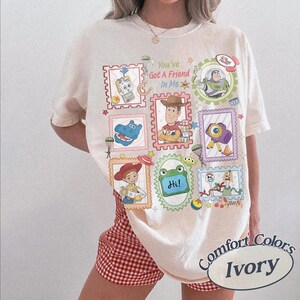 May include: An ivory short-sleeve t-shirt featuring a colorful graphic print of various animated toy characters in postage stamp-style frames. The text "You've Got A Friend In Me" is printed in the center, surrounded by illustrations of characters.