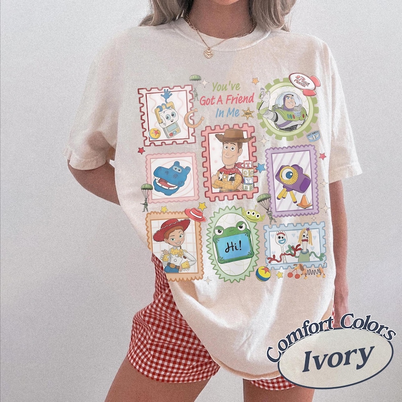 May include: An ivory short-sleeve t-shirt featuring a colorful graphic print of various animated toy characters in postage stamp-style frames. The text "You've Got A Friend In Me" is printed in the center, surrounded by illustrations of characters.