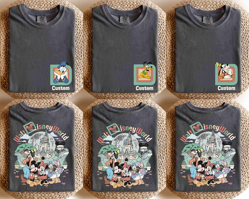 Puede incluir: Seis camisetas de color gris oscuro, dobladas sobre esteras tejidas. Tres camisetas presentan gr&aacute;ficos de personajes de Disney con la palabra "Custom". Las otras tres camisetas muestran un dise&ntilde;o colorido de "Walt Disney World" con personajes de Disney.