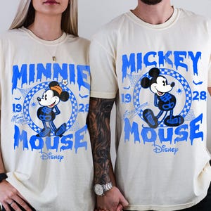May include: Ivory-colored t-shirts featuring blue graphics of Minnie and Mickey Mouse in skeleton costumes. The shirts have the words "Minnie Mouse" and "Mickey Mouse" in a dripping font, with the Disney logo.