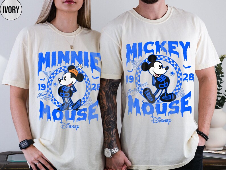 May include: Ivory-colored t-shirts featuring blue graphics of Minnie and Mickey Mouse in skeleton costumes. The shirts have the words "Minnie Mouse" and "Mickey Mouse" in a dripping font, with the Disney logo.