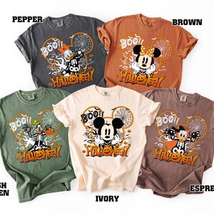 May include: A collection of Halloween-themed t-shirts in various colors, including pepper, brown, ivory, Irish green, and espresso. Each shirt features a Mickey Mouse-shaped design with the word "BOO!!" and Halloween characters.