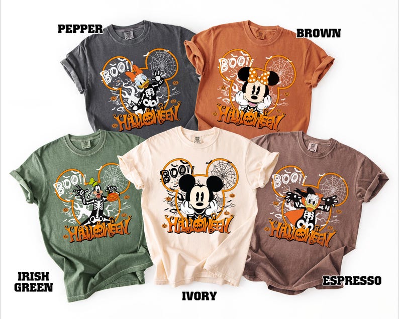 May include: A collection of Halloween-themed t-shirts in various colors, including pepper, brown, ivory, Irish green, and espresso. Each shirt features a Mickey Mouse-shaped design with the word "BOO!!" and Halloween characters.