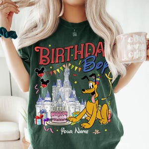 K&ouml;nnte beinhalten: Dunkelgr&uuml;nes T-Shirt mit einer farbenfrohen Grafik mit dem Text "BIRTHDAY Boy" und einer Cartoon-Figur von Pluto. Das Design umfasst ein Schloss, Luftballons, einen Kuchen und die Worte "Your Name".