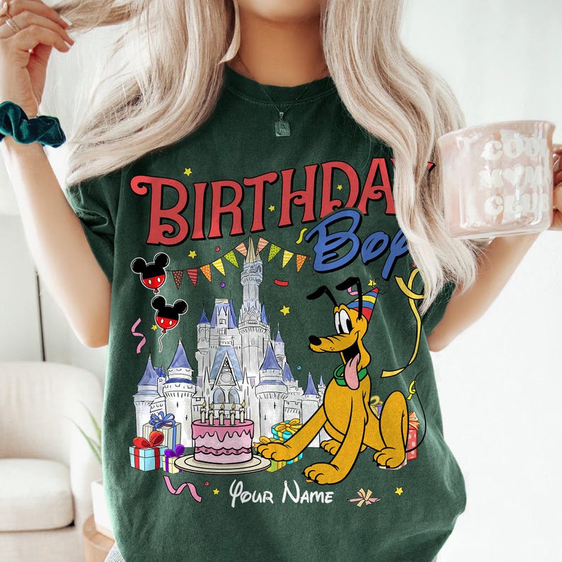 K&ouml;nnte beinhalten: Dunkelgr&uuml;nes T-Shirt mit einer farbenfrohen Grafik mit dem Text "BIRTHDAY Boy" und einer Cartoon-Figur von Pluto. Das Design umfasst ein Schloss, Luftballons, einen Kuchen und die Worte "Your Name".