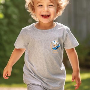 May include: A young child wearing a grey t-shirt with a cartoon character graphic. The character is a blue and white duck wearing a sailor hat. The text "Quackers" is below the character.