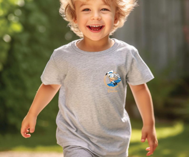 May include: A young child wearing a grey t-shirt with a cartoon character graphic. The character is a blue and white duck wearing a sailor hat. The text "Quackers" is below the character.