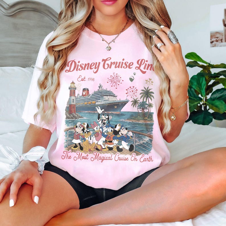 May include: Pale pink t-shirt with a Disney Cruise Line graphic. The design shows a cruise ship, lighthouse, palm trees, fireworks, and Disney characters. Text on the shirt reads "Disney Cruise Line Est. 1998" and "The Most Magical Cruise On Earth."