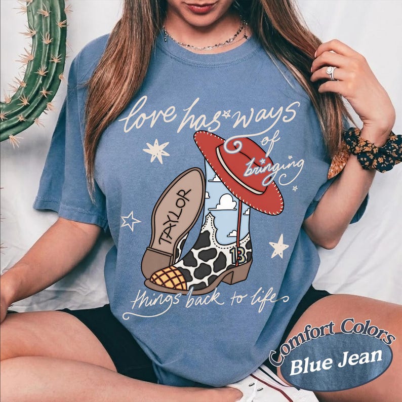 May include: A blue jean colored t-shirt featuring a graphic of a cowboy boot with a cow print pattern, a red hat, and the text "love has ways of bringing things back to life" with "TAYLOR" on the boot and the number "13" on the side.