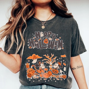 Puede incluir: Camiseta gris oscuro con las palabras "Happy Halloween" en naranja y blanco. La camiseta presenta personajes de dibujos animados, calabazas, fantasmas y otros gr&aacute;ficos tem&aacute;ticos de Halloween. Un dise&ntilde;o divertido y festivo para la temporada de miedo.