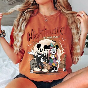May include: Orange t-shirt with a graphic featuring Mickey Mouse and Minnie Mouse dressed as characters from The Nightmare Before Christmas. The text "Nightmare on Main Street" is above the characters.