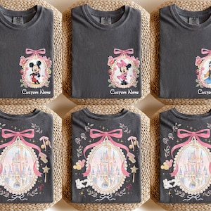 Pode incluir: Seis camisetas cinza carv&atilde;o com diferentes designs. Algumas camisetas apresentam Mickey Mouse, Minnie Mouse e Pateta em molduras ovais com detalhes florais e la&ccedil;os. Outras camisetas exibem um design de castelo com um la&ccedil;o rosa e o texto "Custom Name".