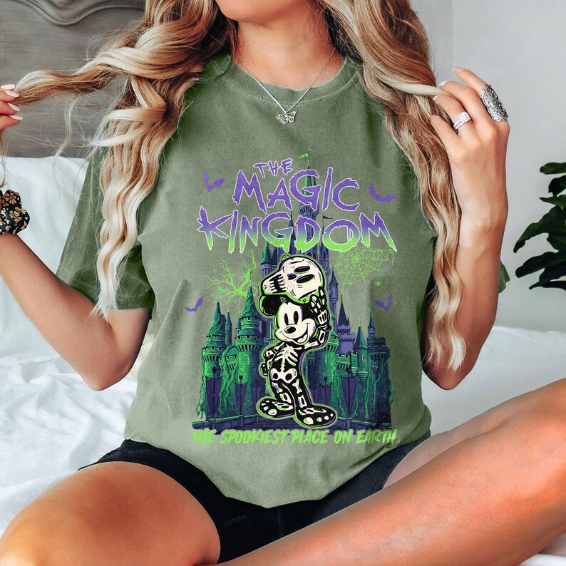 May include: Olive green t-shirt with a Halloween graphic. The design features the text "THE MAGIC KINGDOM" above an illustration of Mickey Mouse in a skeleton costume in front of a castle. The text "THE SPOOKIEST PLACE ON EARTH" is below the castle.