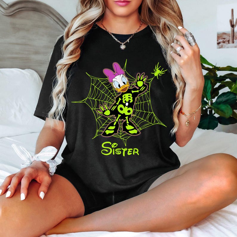 May include: Black t-shirt featuring a Halloween-themed Daisy Duck design. Daisy is depicted as a skeleton within a neon green spiderweb, with the word "Sister" in green script below. A small green spider is also present.