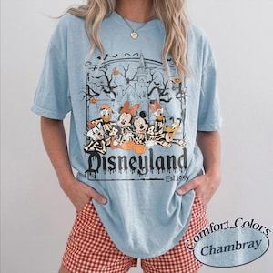 May include: Light blue Disneyland t-shirt featuring a Halloween-themed graphic with Mickey Mouse, Minnie Mouse, Donald Duck, Goofy, and Pluto as skeletons. The shirt has the text "Disneyland Est. 1955". The shirt is made of Comfort Colors Chambray material.