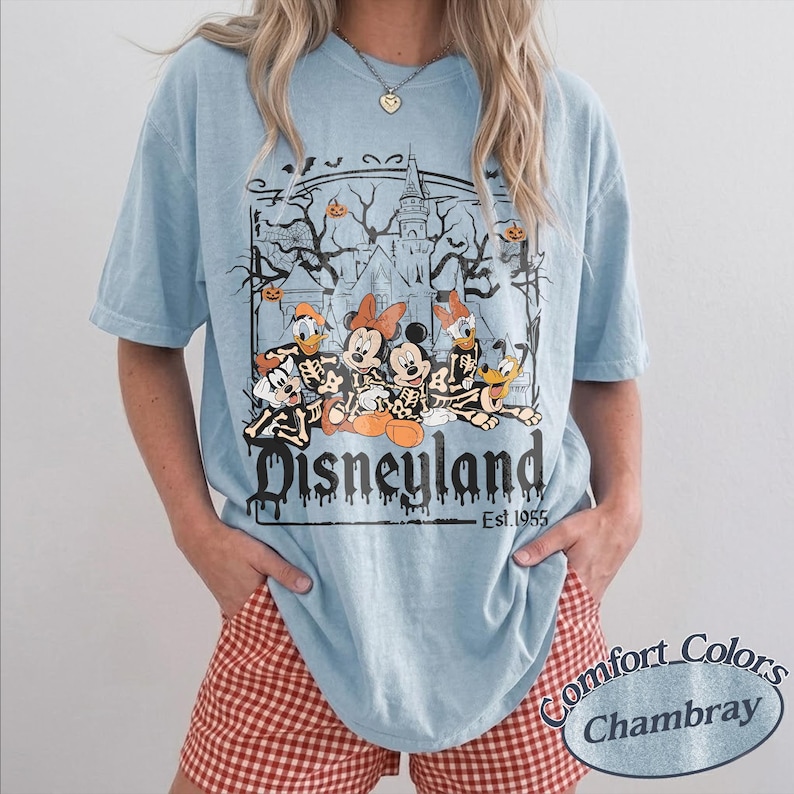 May include: Light blue Disneyland t-shirt featuring a Halloween-themed graphic with Mickey Mouse, Minnie Mouse, Donald Duck, Goofy, and Pluto as skeletons. The shirt has the text "Disneyland Est. 1955". The shirt is made of Comfort Colors Chambray material.