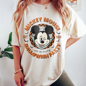 K&ouml;nnte beinhalten: Ein cremefarbenes T-Shirt mit einem Vintage-Mickey-Mouse-Motiv. Das Motiv zeigt Mickey Mouse als Vampir mit dem Text "Mickey Mouse Est 1928 Halloween Party not so scary".