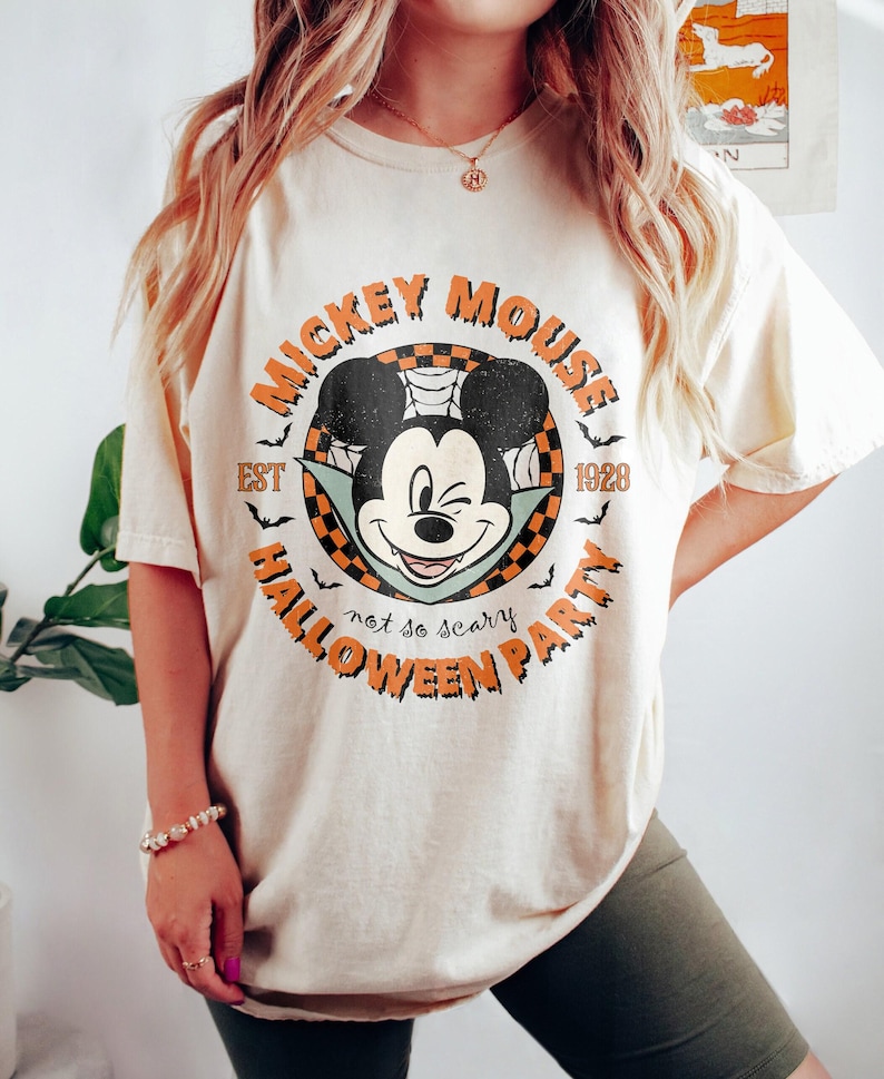 K&ouml;nnte beinhalten: Ein cremefarbenes T-Shirt mit einem Vintage-Mickey-Mouse-Motiv. Das Motiv zeigt Mickey Mouse als Vampir mit dem Text "Mickey Mouse Est 1928 Halloween Party not so scary".