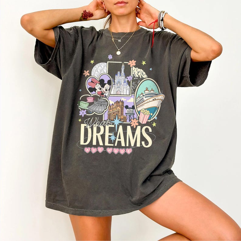 May include: Dark gray oversized t-shirt featuring a colorful graphic with the text "What Dreams". The graphic includes images of a cruise ship, a castle, and cartoon characters. The shirt has short sleeves and a relaxed fit.