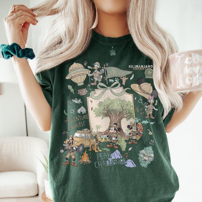 May include: Dark green t-shirt featuring a safari-themed graphic with Mickey and Minnie Mouse, Donald Duck, and Goofy. The design includes a hot air balloon, a jeep, and the text "Kilimanjaro Safari" and "Adventure Awaits!"