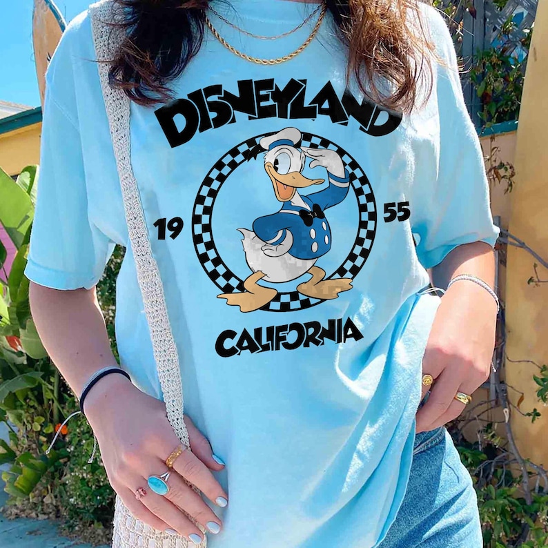 K&ouml;nnte beinhalten: Hellblaues T-Shirt mit einer Grafik von Donald Duck in Matrosenkleidung, mit dem Text "Disneyland California 1955". Das Design befindet sich in einem karierten Kreis. Das Shirt hat kurze &Auml;rmel und einen Rundhalsausschnitt.