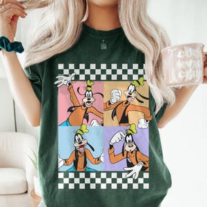 May include: Dark green t-shirt featuring a four-panel graphic of Goofy in various poses. The graphic is framed by a black and white checkered border. Goofy is wearing an orange outfit with a black vest and a green hat.