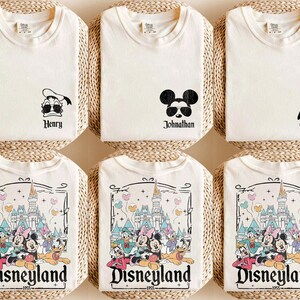 K&ouml;nnte beinhalten: Sechs wei&szlig;e T-Shirts mit verschiedenen Designs. Drei Shirts zeigen eine Comicfigur: Donald Duck, Mickey Mouse und Goofy. Die anderen drei Shirts zeigen ein Comicbild von Disneyland mit dem Text "Disneyland 1955".