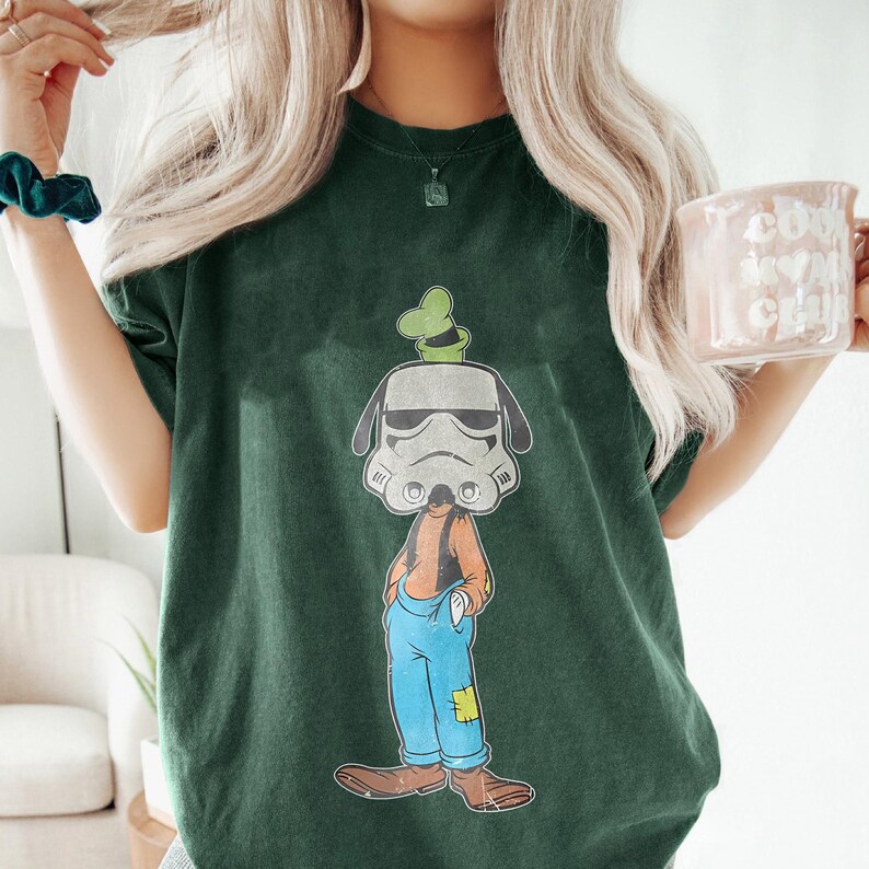 May include: Dark green t-shirt featuring a cartoon character with a Stormtrooper helmet, wearing blue overalls and brown shoes. The character has a green hat. The shirt is a relaxed fit.