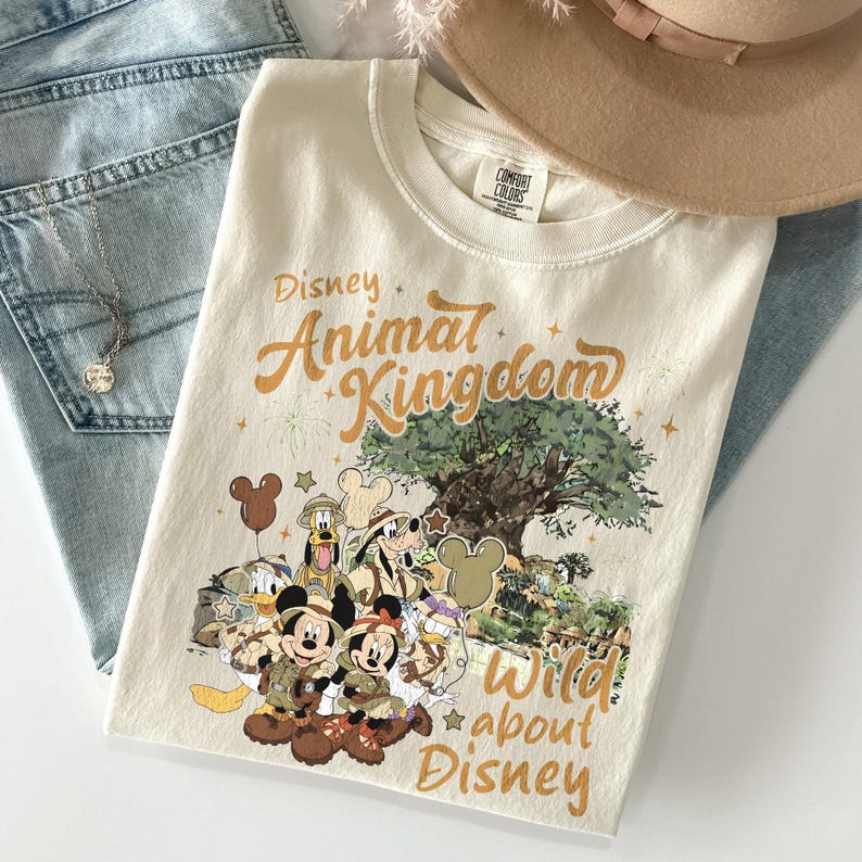 Pu&ograve; includere: T-shirt color crema con la scritta "Disney Animal Kingdom" in lettere dorate, con una grafica di personaggi Disney e un albero. La maglietta &egrave; piegata sopra jeans blu, con una collana e un cappello beige visibili.
