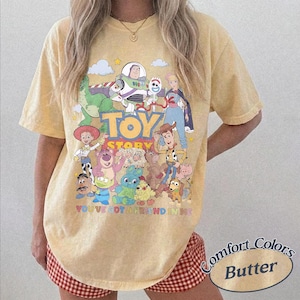 May include: A pale yellow short-sleeve t-shirt featuring a colorful graphic print of various Toy Story characters, the Toy Story logo, and the text You've Got A Friend In Me. The shirt has a crew neckline and a relaxed, loose-fitting silhouette.