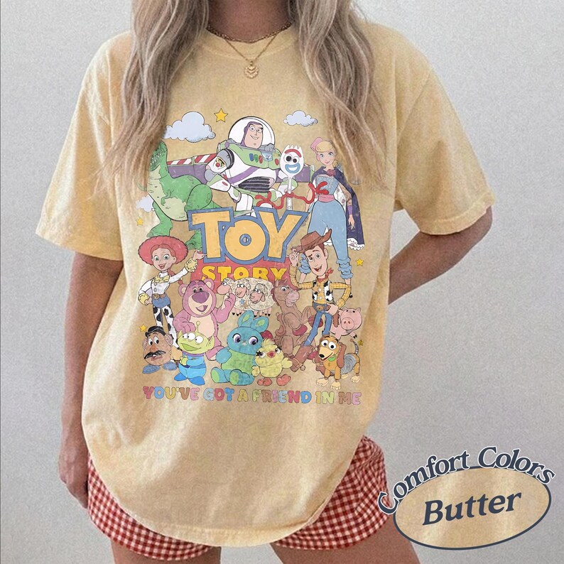 May include: A pale yellow short-sleeve t-shirt featuring a colorful graphic print of various Toy Story characters, the Toy Story logo, and the text You've Got A Friend In Me. The shirt has a crew neckline and a relaxed, loose-fitting silhouette.