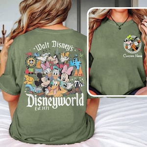 May include: Olive green t-shirt featuring a colorful graphic of Disney characters and the text "Walt Disney's Disneyworld Est. 1971" on the back. A smaller image of Goofy and Pluto is on the front, with space for a custom name.
