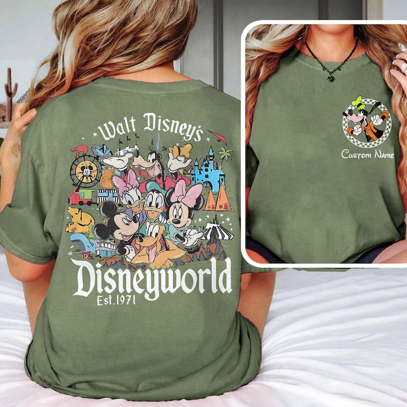 May include: Olive green t-shirt featuring a colorful graphic of Disney characters and the text "Walt Disney's Disneyworld Est. 1971" on the back. A smaller image of Goofy and Pluto is on the front, with space for a custom name.
