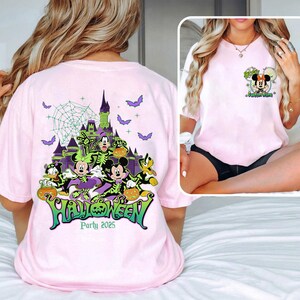 Op de afbeelding: Lichtroze t-shirt met een Halloween-ontwerp. Het ontwerp toont Disney-figuren, een spookkasteel en vleermuizen. Het woord "Halloween" is groen, met de tekst "Party 2025".
