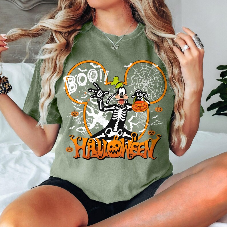 May include: Olive green t-shirt featuring a Halloween-themed design. The graphic includes a skeleton character with a pumpkin, the word "BOO!!", and the word "HALLOWEEN" in orange. The design is set within a Mickey Mouse head shape.